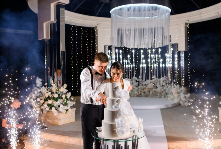 front-view-happy-married-couple-loving-man-and-woman-cutting-big-white-cake-on-wedding-ceremony-at-night-standing-near-welldecorated-luxury-altar-with-flowers-and-sparklers-special-event